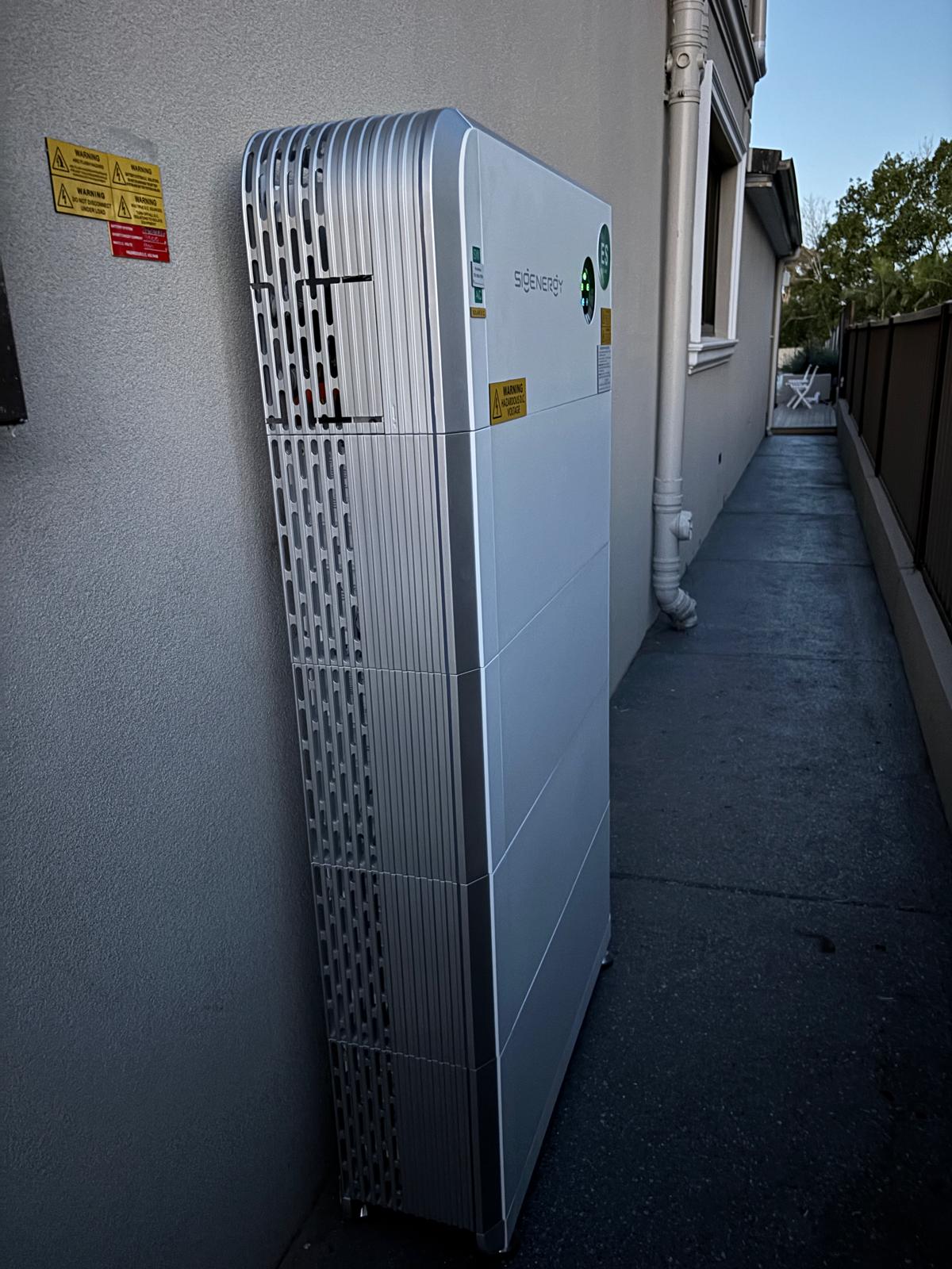 Sigenergy SigenStor battery installed along a side path, cabling and warning labels in place.