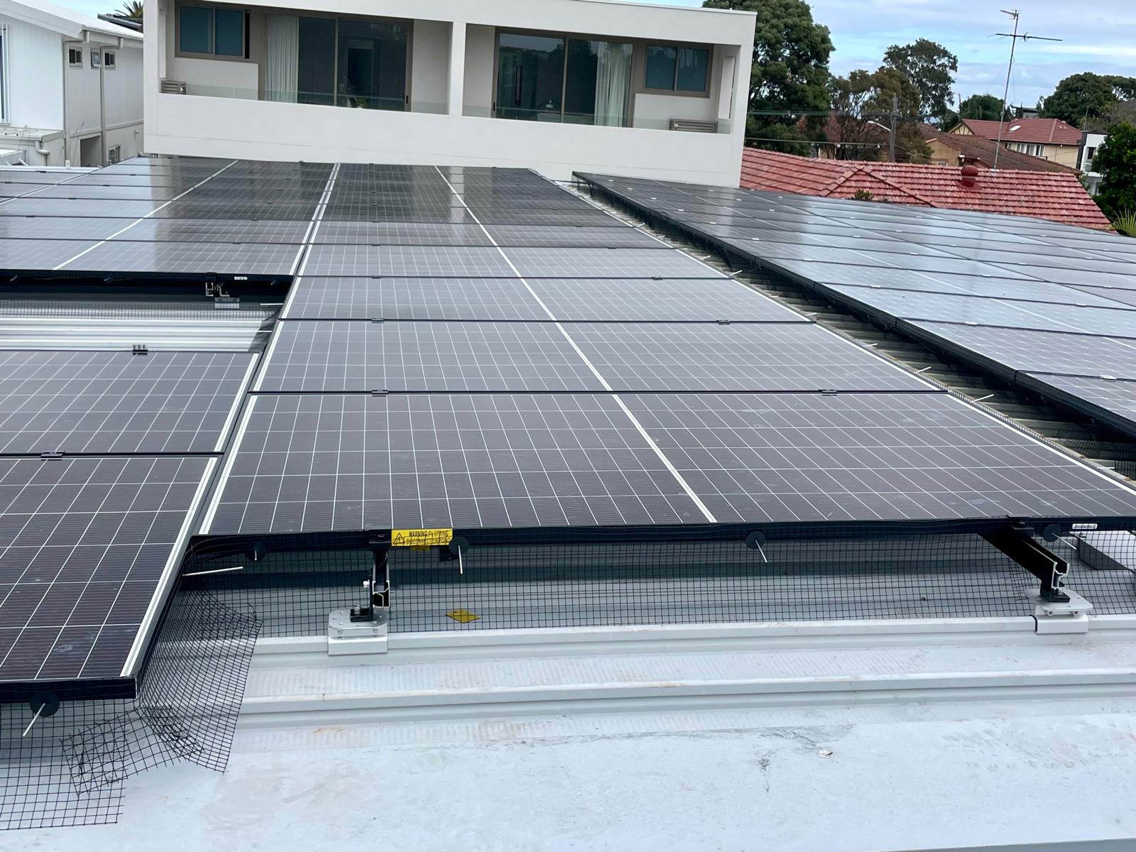 Solar panels on tilt frames over a low-pitch metal roof, edged with bird-proofing mesh.